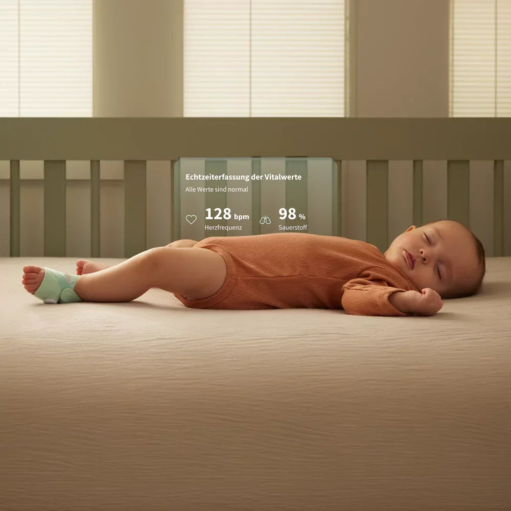 Ein Baby schläft friedlich in einem braunen Strampler auf einem Bett und trägt ein kleines Gerät an einem Fuß. Im Hintergrund zeigt ein Display die Herzfrequenz (128 Schläge/min) und die Sauerstoffsättigung (98 %) an.