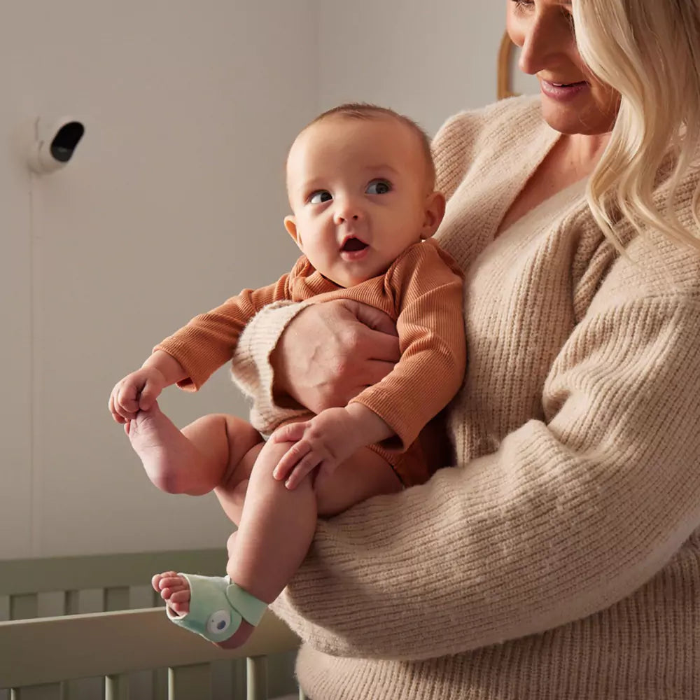 Eine Frau mit langen blonden Haaren und einem beigen Pullover hält ein Baby in einem rostfarbenen Outfit. Das Baby trägt eine grüne Socke an einem Fuß und scheint zur Seite zu schauen. Im Hintergrund sind ein Kinderbett und ein an der Wand montiertes Babyphone zu sehen.
