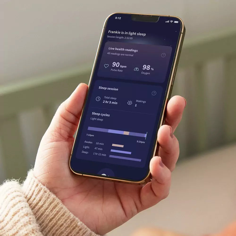 Eine Person hält ein Smartphone mit einer Schlafüberwachungs-App, die einem modernen Babyphone ähnelt. Die App zeigt Live-Gesundheitswerte mit einer Pulsfrequenz von 90 Schlägen pro Minute und einem Sauerstoffgehalt von 98 %. Sie liefert auch Details zu Schlafsitzungen und -zyklen. Der Bildschirm des Telefons ist dunkel mit weißem Text.