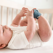 Ein Baby liegt auf dem Rücken in einem Kinderbett und trägt einen weißen Strampler und blaue Stoffschuhe. Es umfasst sanft seine Füße mit beiden Händen und blickt mit ruhigem Gesichtsausdruck nach oben. Eine Owlet Dream Sock: Medizinisch zertifiziertes Pulsoximeter für Babys von Owletcare.de ist an einem winzigen Finger befestigt und sorgt für zusätzliche Beruhigung in der sanft konzentrierten, heiteren Atmosphäre.