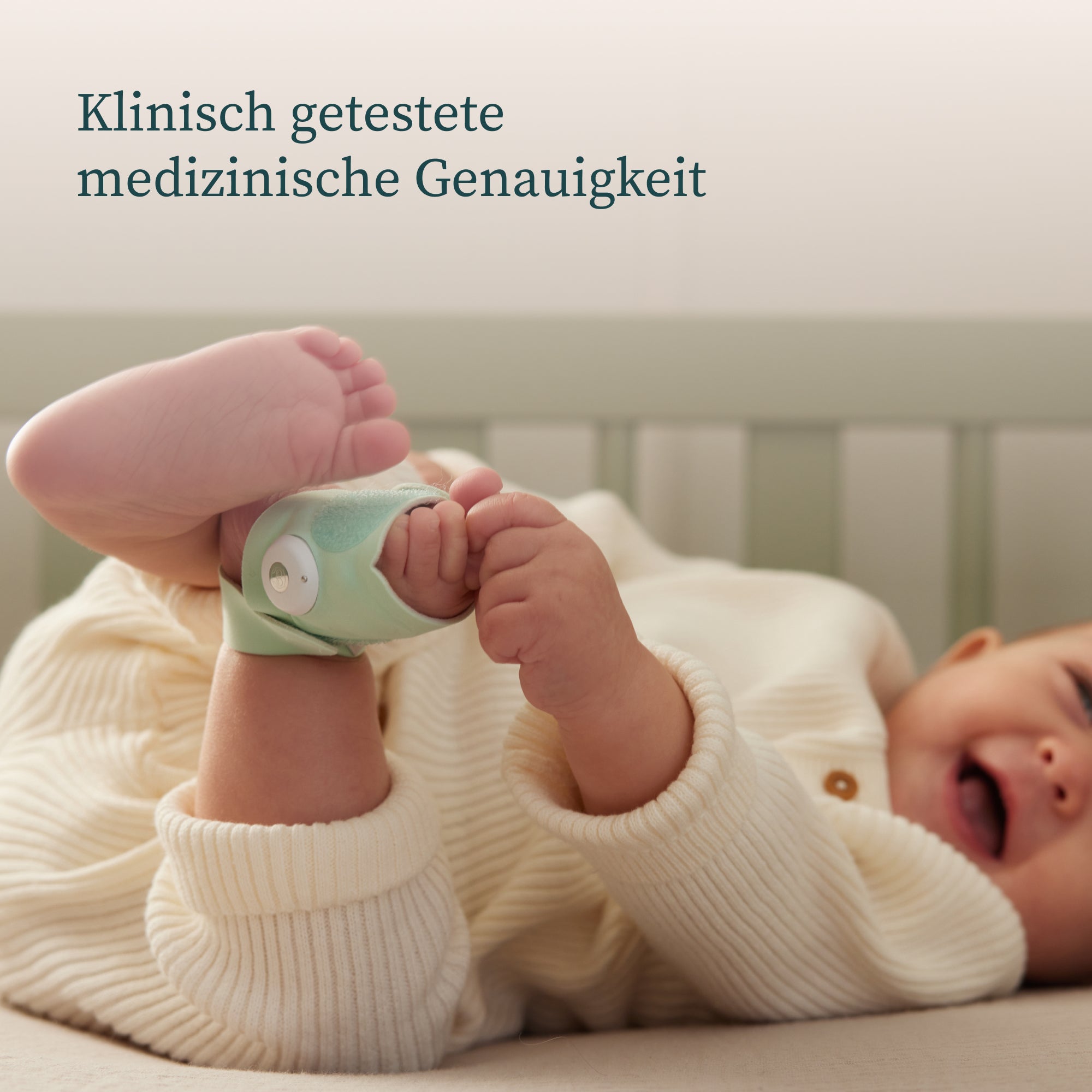 Ein Baby in einem weißen Strick-Strampler liegt auf dem Rücken in einem Kinderbett und spielt mit seinen Füßen. An einem Fuß ist eine kleine grüne Owlet Dream Sock befestigt: Medizinisch zertifiziertes Pulsoximeter für Babys. Der deutsche Text oben lautet „Klinisch getestete medizinische Genauigkeit“. Das Produkt ist bei Owletcare.de erhältlich.