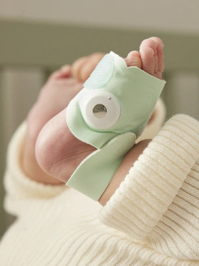 Nahaufnahme eines Babyfußes, der mit einem tragbaren Babypulsoximeter geschmückt ist, das in weichen mintgrünen Stoff eingewickelt ist und an dessen Oberseite sich ein kleiner runder Sensor befindet. Das Baby ist in ein cremefarbenes Strickoutfit gekleidet. Der Hintergrund ist unscharf, scheint aber ein Kinderbett zu zeigen.