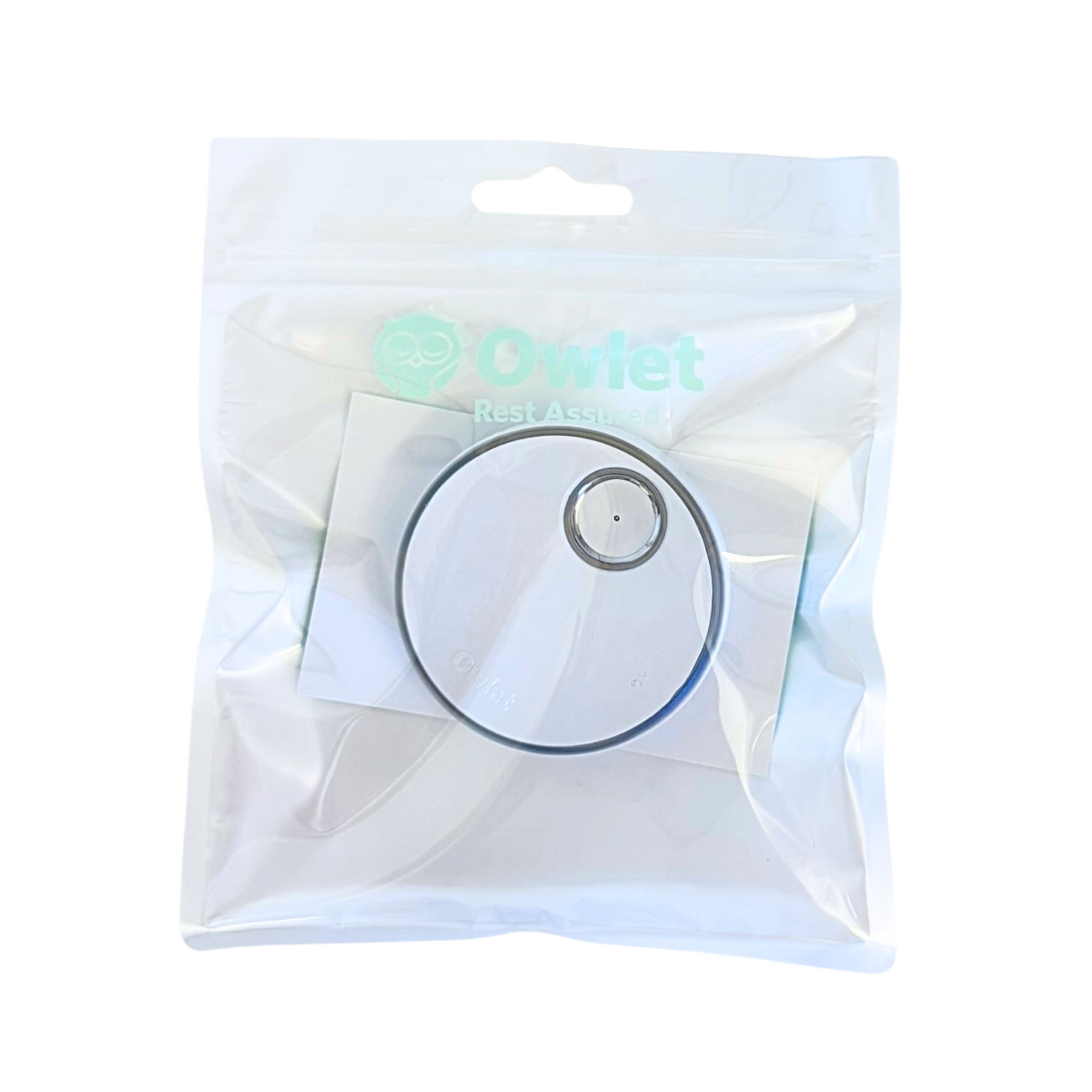 In einer versiegelten, transparenten Plastiktüte befindet sich die Owlet Dream Sock Basisstation mit einem kreisförmigen Sensor und dem Owlet-Logo mit dem Text "Rest Assured" auf der Verpackung.