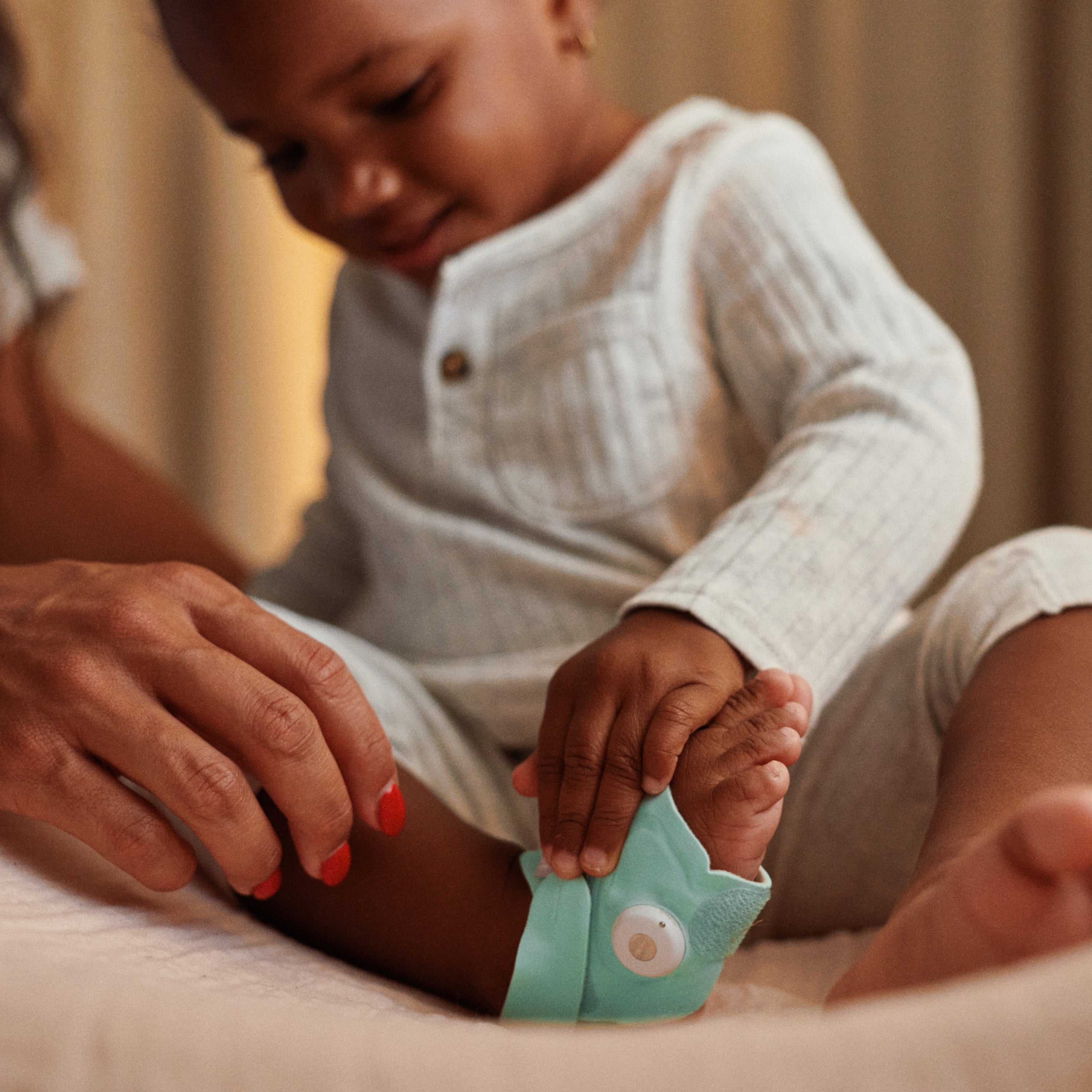 Ein Kleinkind in einem hellen Schlafanzug sitzt auf einem Bett, während ein Erwachsener dabei hilft, das „Owlet Dream Duo mit Ersatzsocke“ von Owletcare.de – ein türkisfarbenes tragbares Überwachungsgerät – am Fuß des Kindes anzulegen.