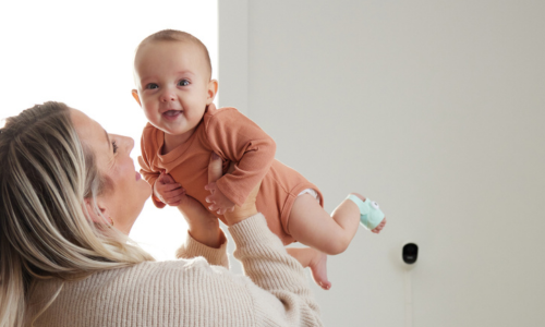 Eine Frau lächelt und hält ein glückliches Baby in einem orangefarbenen Outfit in die Luft. Das Baby, das an einem Fuß ein Baby-Pulsoximeter trägt, strahlt vor Freude. Im Hintergrund ist an einer weißen Wand ein Babyüberwachungsgerät montiert.