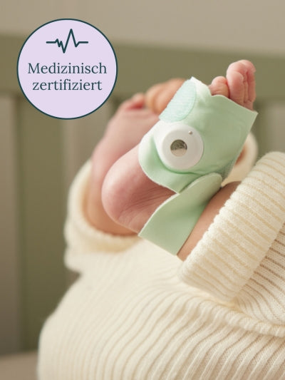 Eine Nahaufnahme des Fußes eines Babys, der ein hellgrünes Baby-Pulsoximeter trägt. Der Fuß, der in ein weißes Strickoutfit gehüllt ist, wird sanft angehoben. In der oberen linken Ecke befindet sich ein rundes Zertifizierungsabzeichen mit einem Herzmonitorsymbol und dem Text „Medizinisch zertifiziert“.