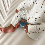 Man sieht den Unterkörper und die Hände eines Babys, das auf einer Matratze sitzt und einen weißen Strampler mit braunen Punkten und eine blaue Owletcare.de Traumsocke aus dem Owlet Dream Bundle trägt: Traumsocke + Cam 2.