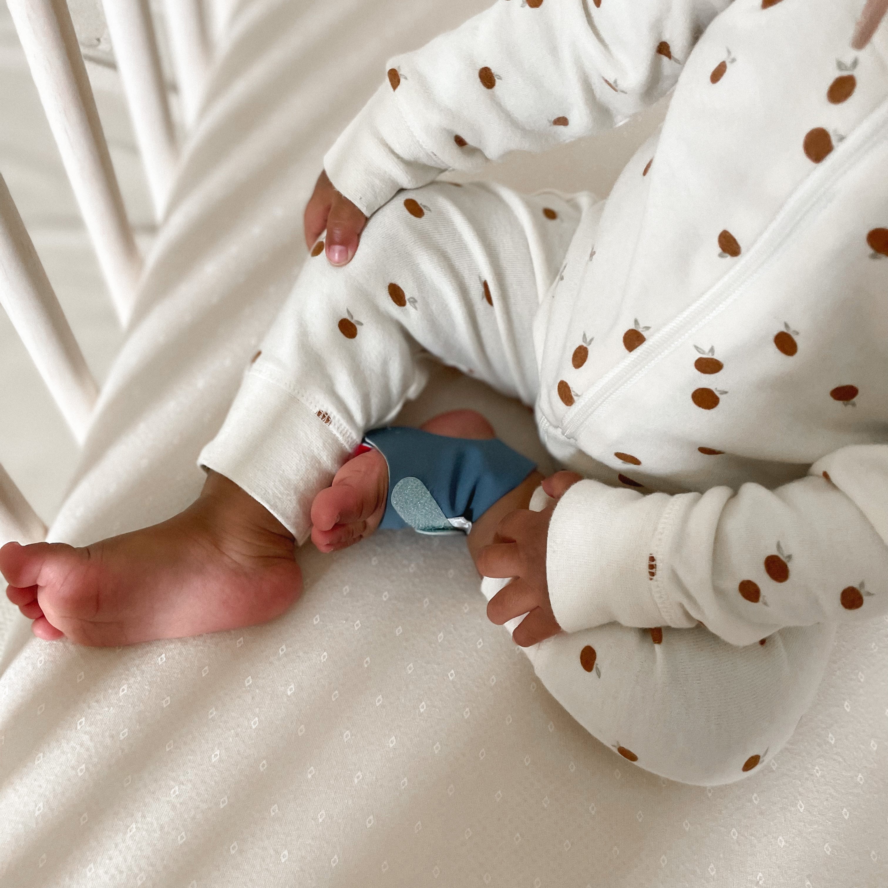 Man sieht den Unterkörper und die Hände eines Babys, das auf einer Matratze sitzt und einen weißen Strampler mit braunen Punkten und eine blaue Owletcare.de Traumsocke aus dem Owlet Dream Bundle trägt: Traumsocke + Cam 2.