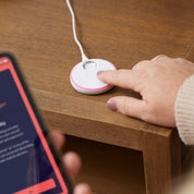 Eine Person drückt eine kleine, runde, weiße Owlet Dream Sock aus dem Owletcare.de Dream Bundle: Dream Sock + Cam 2 auf eine hölzerne Oberfläche, während sie ein Smartphone hält, auf dem die Babyüberwachungs-App im Vordergrund zu sehen ist.