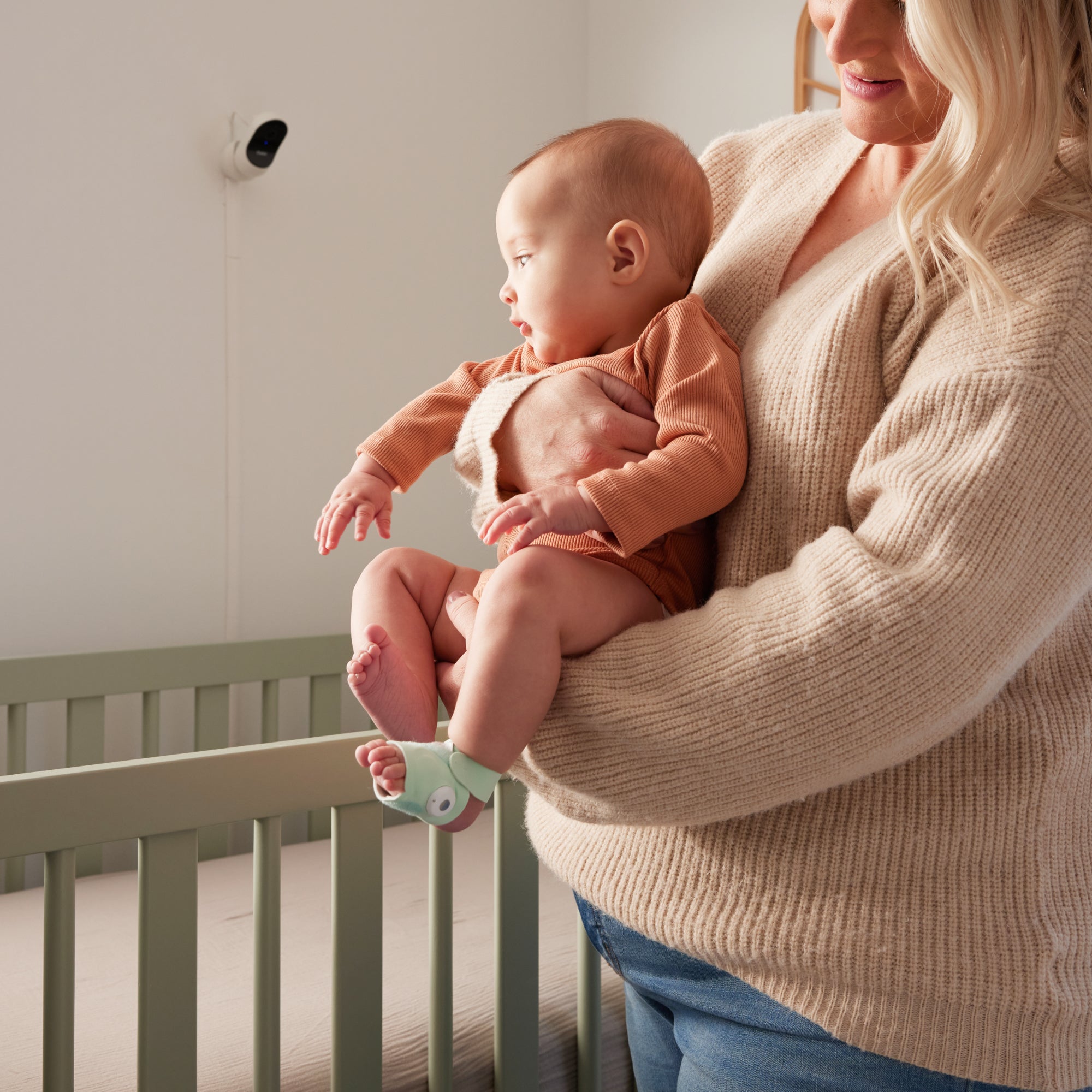 Eine Frau steht neben einem hellgrünen Kinderbett in einem sanft beleuchteten Kinderzimmer und hält ein Baby in Braun, das die mintgrüne Owletcare.de Traumsocke aus dem Owlet Dream Bundle trägt: Traumsocke + Cam 2.