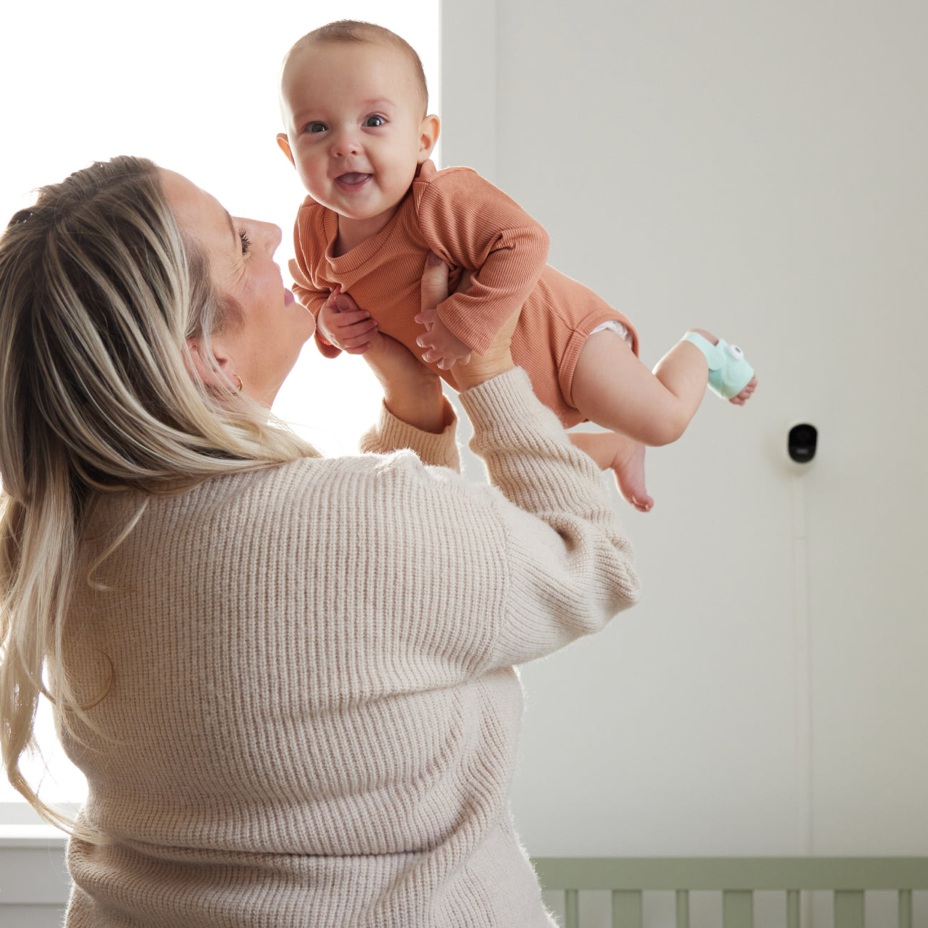 Eine Frau hebt ein lächelndes Baby in einem braunen Strampler hoch, das die Owletcare.de Traumsocke aus dem Owlet Dream Bundle trägt: Traumsocke + Cam 2. Neben ihnen steht ein Kinderbett und die Cam 2 WiFi-Kamera, die wertvolle Momente in Innenräumen festhält.