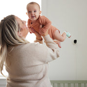 Eine Frau hebt ein lächelndes Baby in einem braunen Strampler hoch, das die Owletcare.de Traumsocke aus dem Owlet Dream Bundle trägt: Traumsocke + Cam 2. Neben ihnen steht ein Kinderbett und die Cam 2 WiFi-Kamera, die wertvolle Momente in Innenräumen festhält.