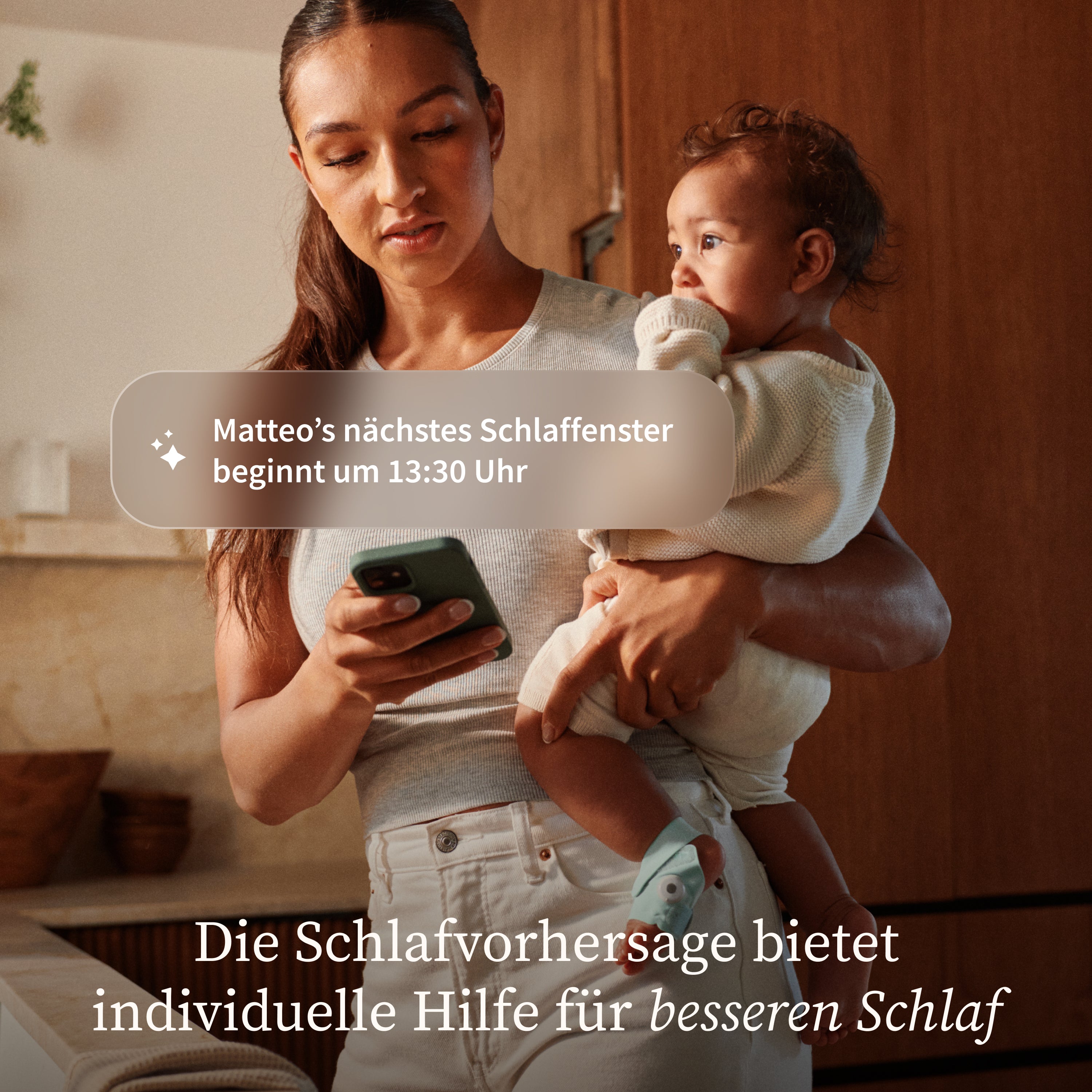 Eine Frau mit einem Baby im Arm schaut in der Küche auf ihr Smartphone. Der Text auf dem Bildschirm informiert über Matteos nächstes Schlaffenster um 13:30 Uhr und hebt hervor, dass Owletcare.de's Owlet Dream Duo - Dream Sock + Dream Sight Kamera - eine persönliche Schlafanalyse für besseren Schlaf bietet.