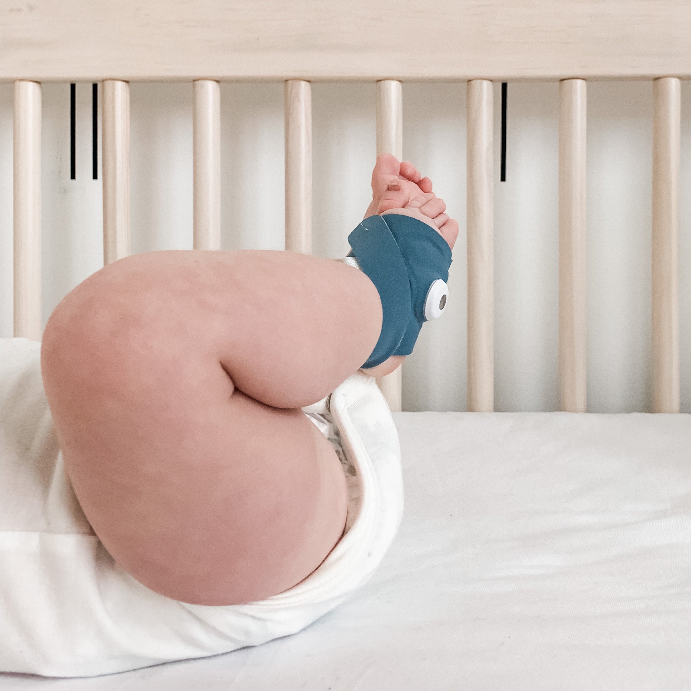 Ein Baby liegt auf einem weißen Laken in einem Kinderbett, wobei nur seine Beine und der Unterkörper zu sehen sind; an einem Fuß trägt es die „Owlet Dream Sock“ mit Ersatzsocke von Owletcare.de.