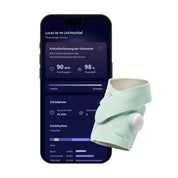 Auf einem Smartphone wird eine Schlaf-Tracking-App mit Herzfrequenz- und Sauerstoffdaten in Echtzeitüberwachung angezeigt. Vor dem Smartphone liegt der „Owlet Dream Sock mit Ersatzsocke“ von Owletcare.de, ein hellgrünes, weiches Armband mit einem runden Sensor.