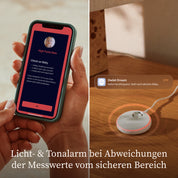 Eine Person hält ein Smartphone in der Hand, auf dem eine Warnmeldung wegen eines hohen Pulswerts ihres Babys angezeigt wird, während die „Owlet Dream Sock“ mit Ersatzsocke von Owletcare.de mit ihrem leuchtenden Rand auf einer Holzunterlage liegt. Unten ist folgender deutscher Text zu lesen: „Licht- und Tonalarm bei Abweichungen der Messwerte vom sicheren Bereich.“.