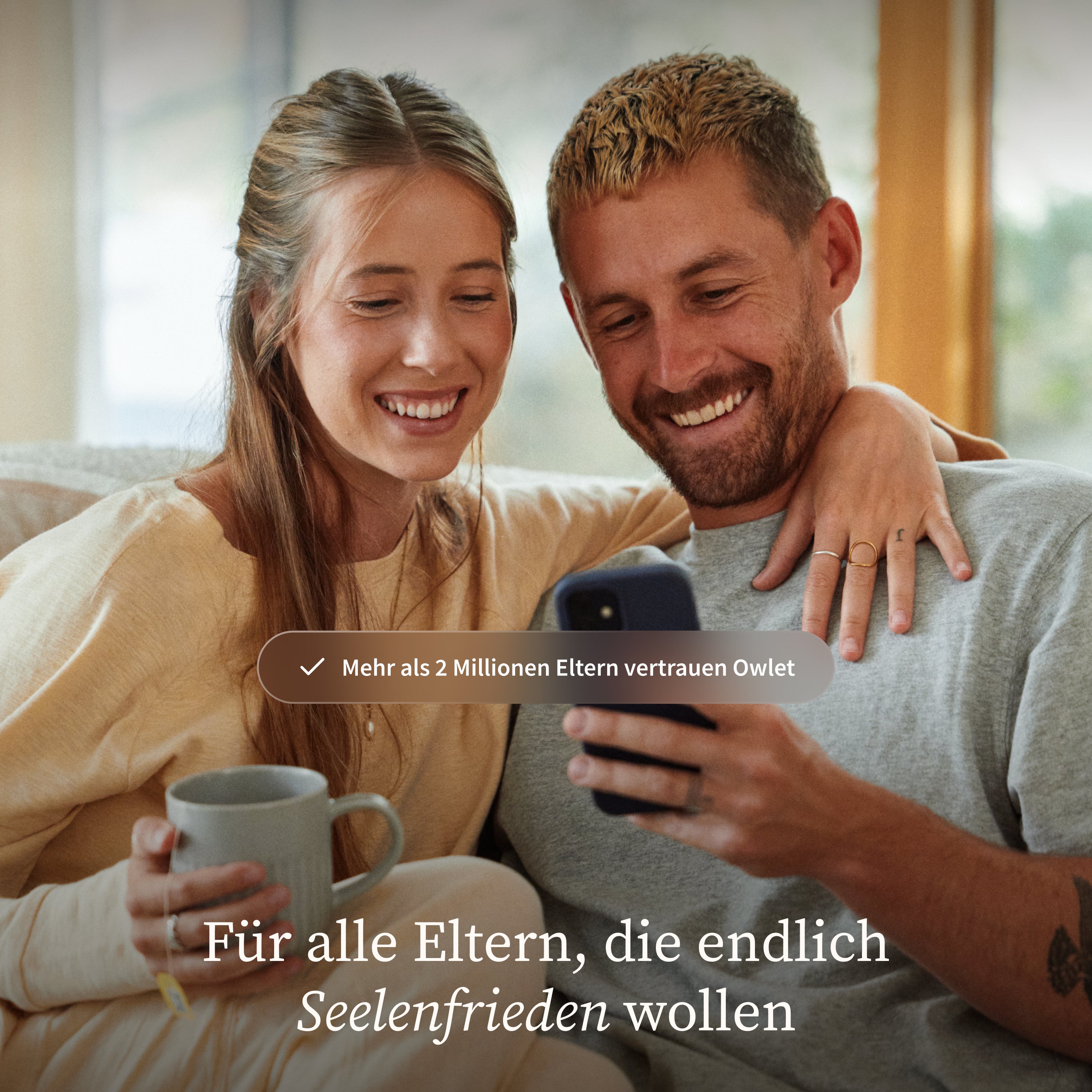 Ein lächelndes Paar sitzt gemeinsam auf einem Sofa und schaut auf ein Smartphone. Die Frau hält eine Tasse in der Hand, während ein deutscher Text das Vertrauen in den „Owlet Dream Sock mit Ersatzsocke“ von Owletcare.de weckt, der Eltern ein beruhigendes Gefühl gibt.