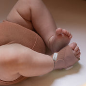Eine Nahaufnahme eines Babys, das braune, gerippte Shorts und an einem Fuß die „Owlet Dream Sock mit Ersatzsocke“ von Owletcare.de trägt und auf einem hellen Untergrund liegt. Die Beine und Füße des Babys sind entspannt und leicht angewinkelt.