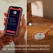Eine Person hält ein Smartphone in der Hand, auf dem eine Warnmeldung von Owletcare.de wegen einer hohen Pulsfrequenz des Owlet Dream Duo mit Ersatzsocke angezeigt wird. Daneben befindet sich das runde Sensorgerät mit einem leuchtenden Rand. Der Text darunter lautet: „Licht- und Tonalarm bei Abweichungen der Messwerte vom sicheren Bereich.“.