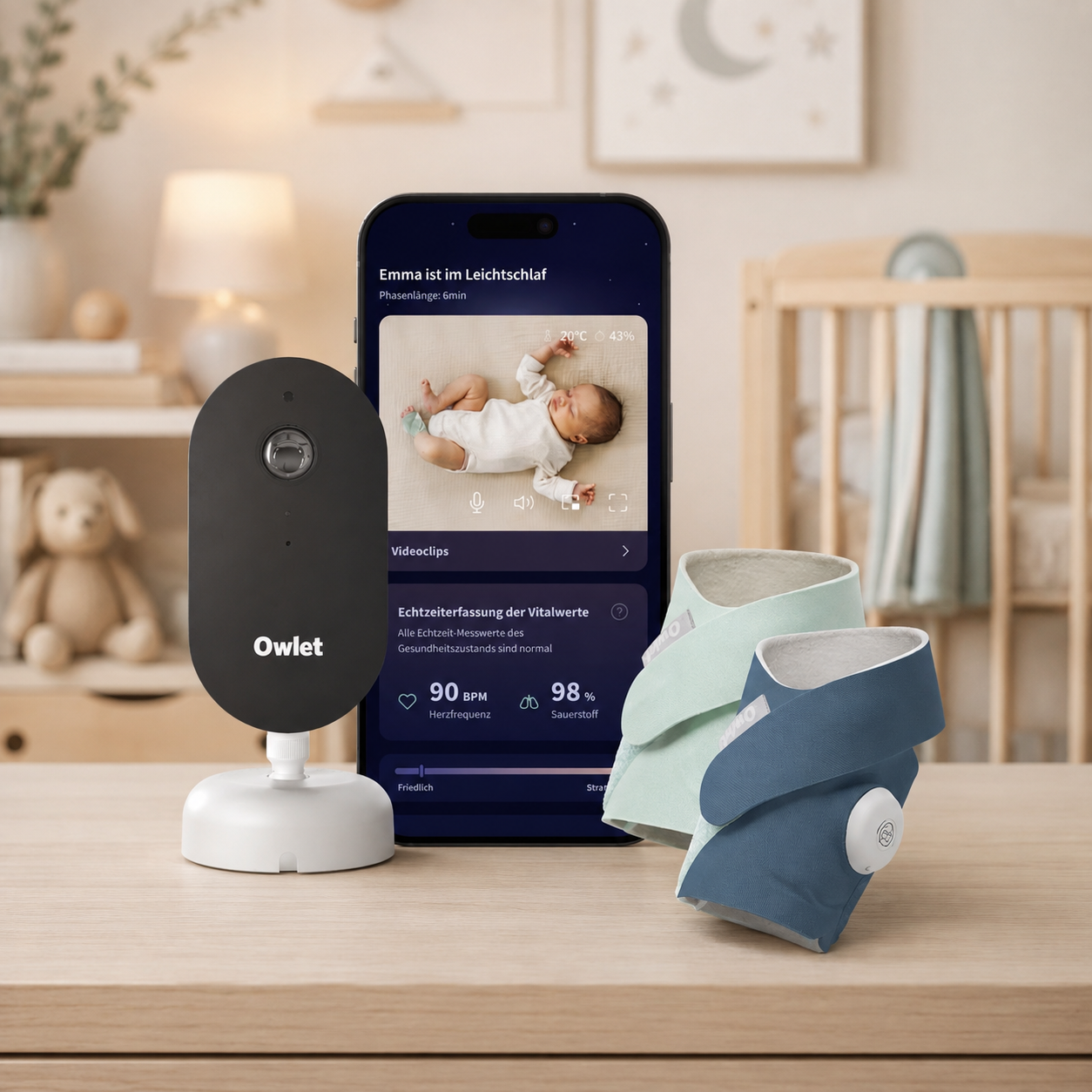 Ein „Owlet Dream Duo“ von Owletcare.de mit Ersatzsöckchen – bestehend aus einer Babyphone-Kamera, zwei tragbaren Babysöckchen und einem Smartphone, auf dem die App für Schlaf- und Vitaldaten angezeigt wird – steht auf einem Holztisch in einem sanft beleuchteten Kinderzimmer mit einem Kinderbett, Plüschtieren und einer neutralen Einrichtung.