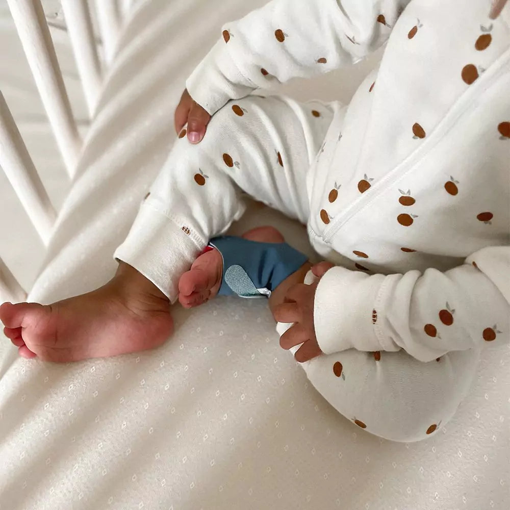 Ein Baby im weißen Pyjama mit braunen Punkten sitzt auf einem hellen Laken. Ein Teil des Gitters des Kinderbetts ist zu sehen. Am nackten Fuß des Babys ist eine Owletcare.de Owlet Dream Sock: Medizinisch zertifiziertes Pulsoximeter für Babys befestigt.